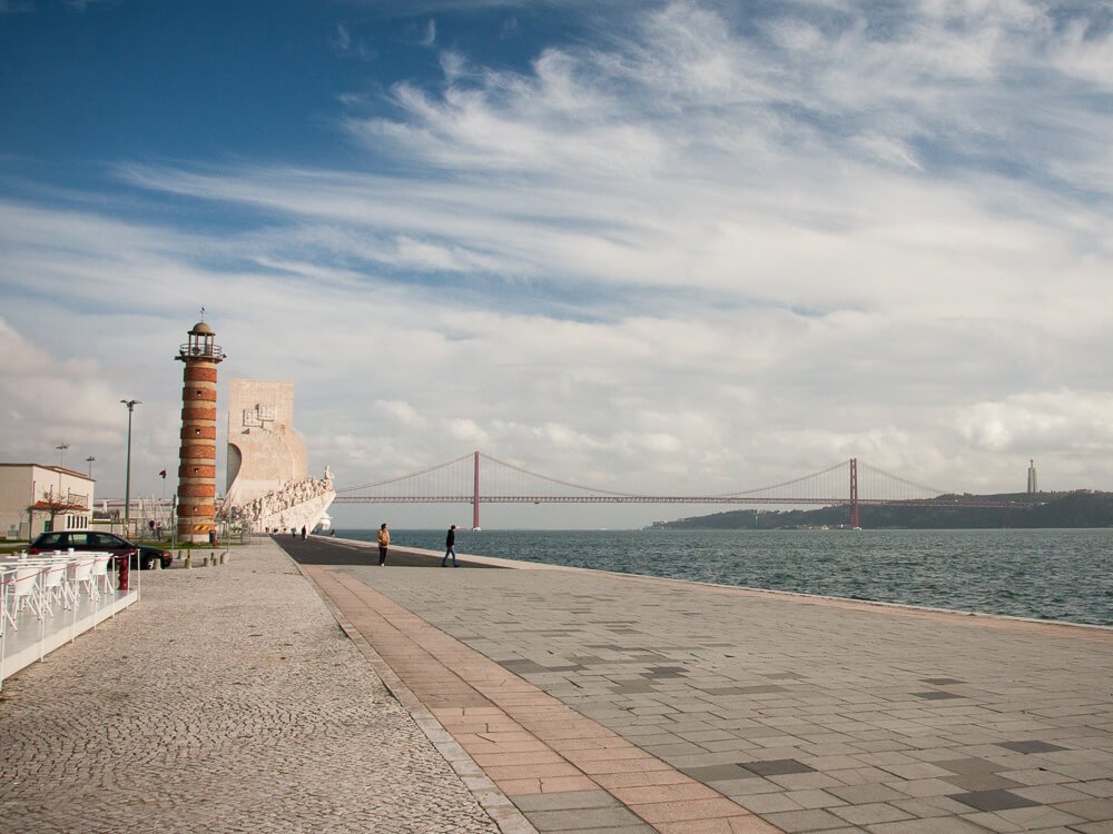 A view of the Monument to the Discoveries, a red lighthouse, and the 25 de Abril Bridge along the Tagus River in Lisbon.