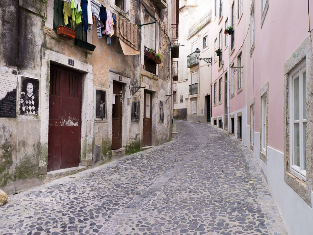 A narrow cobblestone street lined with old buildings, some with laundry hanging outside, in a Lisbon neighborhood.