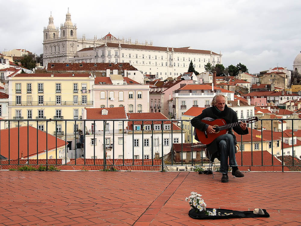 A street musician playing guitar at a viewpoint overlooking the Alfama neighborhood, with the Church of São Vicente de Fora in the background.