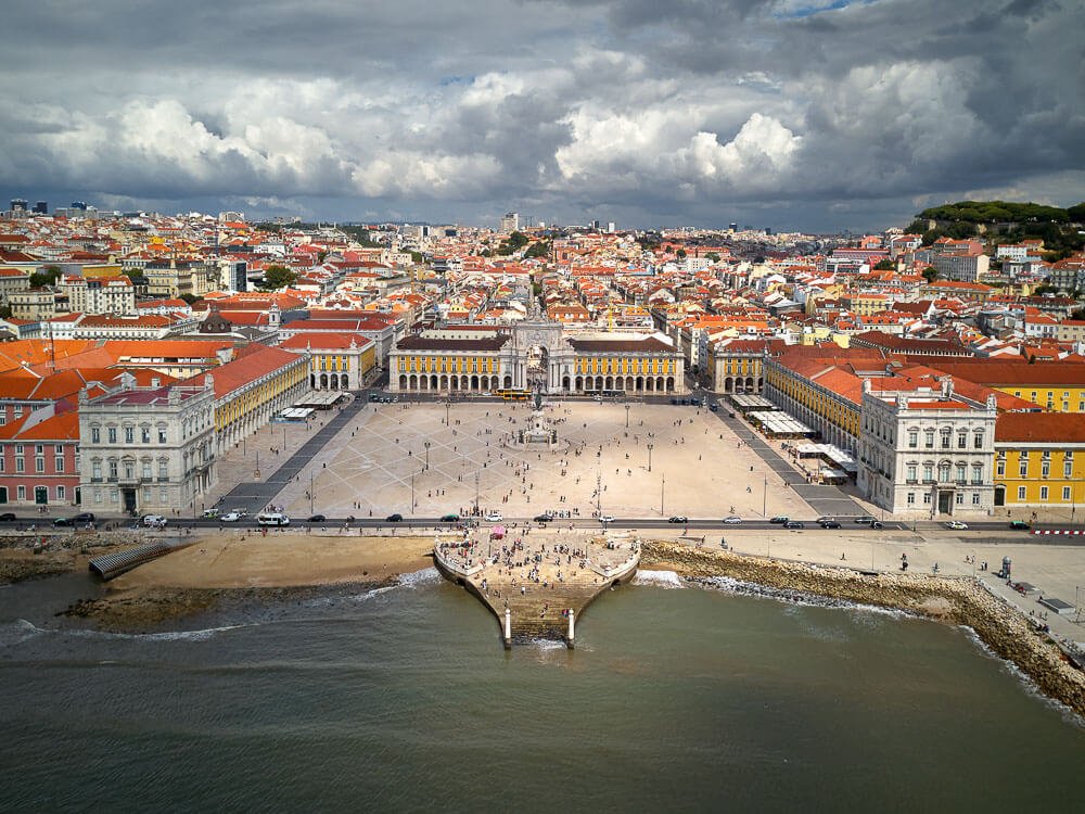 An aerial view of Praça do Comércio, a large square in Lisbon, with surrounding historic buildings and the Tagus River in the background.