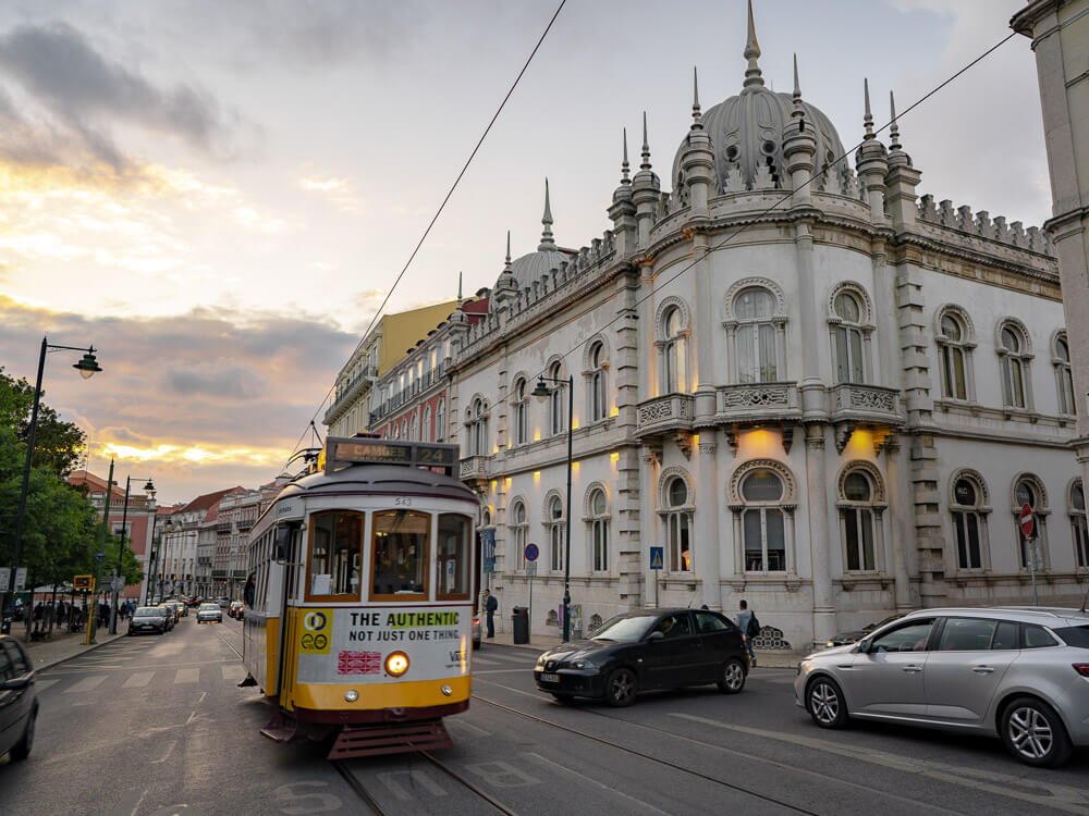 A classic yellow tram driving past a historic building in Lisbon at sunset, with the sky showing hues of orange and pink.