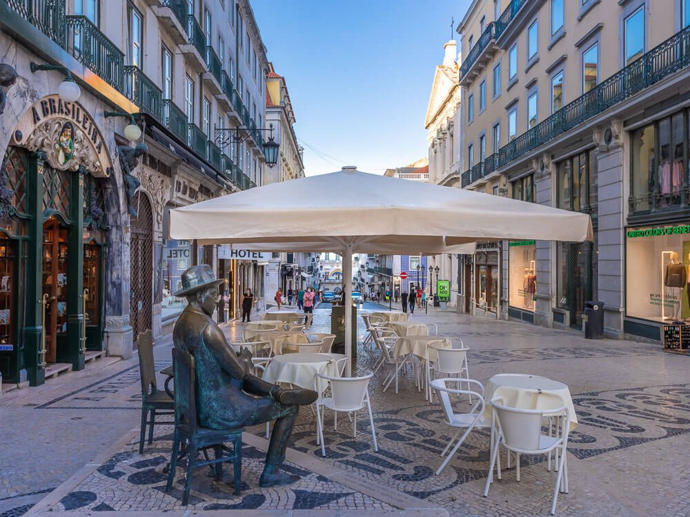 A café terrace with white tables and chairs, and a bronze statue of Fernando Pessoa, outside A Brasileira café in Lisbon.