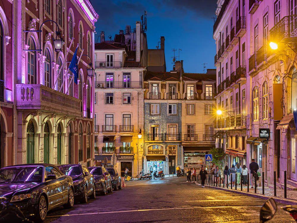 A Lisbon street at night with buildings illuminated in purple and yellow lights, with people walking along the sidewalks.