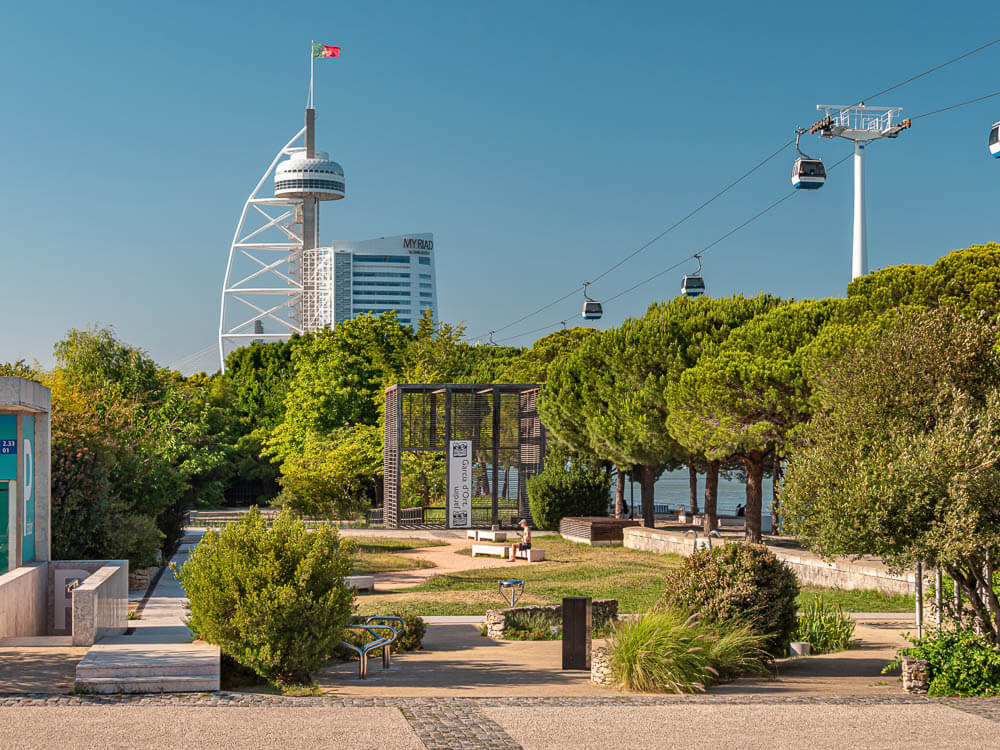 A green park area with trees and a modern tower in the background, along with cable cars in the Parque das Nações area of Lisbon.