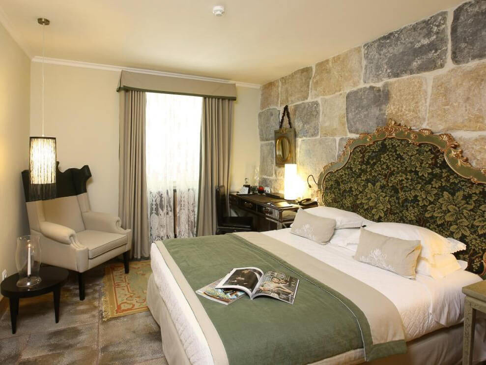 Cozy boutique hotel room with a green and gold ornate headboard, stone accent wall, and a comfortable armchair in Lisbon.