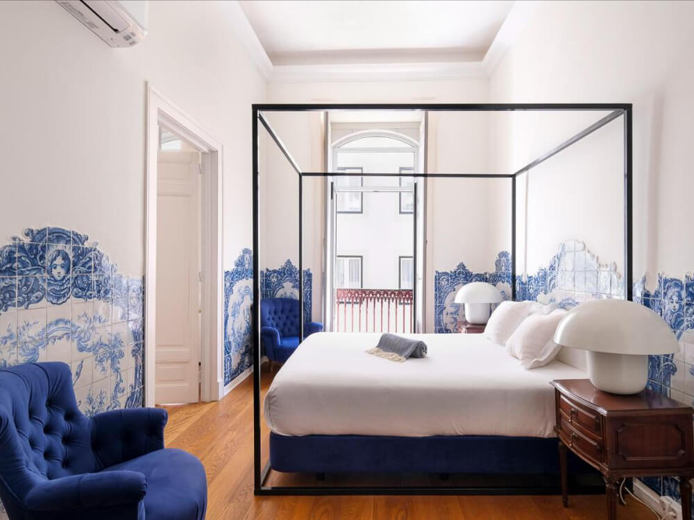 Elegant hotel room with a four-poster bed, blue and white tile details on the walls, and matching blue armchairs.