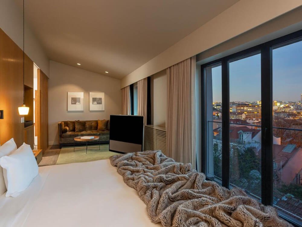 Chic hotel room with a large bed, a fur throw, and floor-to-ceiling windows offering a stunning city view.