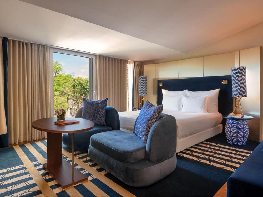 Elegant hotel room with plush blue seating, a stylish bed, and large windows providing a view of the surrounding greenery.