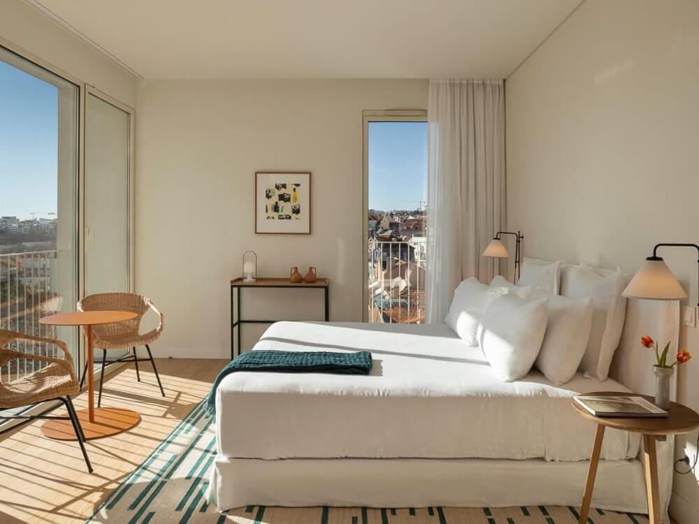 Sunlit boutique hotel room with large windows, simple decor, and a balcony with city views in Lisbon.